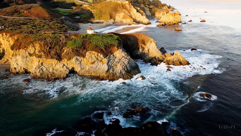 Drone view of waves crashing against the rugged cliffs along Big Sur’s garay point beach. The crashing blue waters create white-tipped waves, while the golden light of the setting sun illuminates the rocky shore. A small island with a lighthouse sits in the distance, and green shrubbery covers the cliff’s edge. The steep drop from the road down to the beach is a dramatic feat, with the cliff’s edges jutting out over the sea. This is a view that captures the raw beauty of the coast and the rugged landscape of the Pacific Coast Highway.