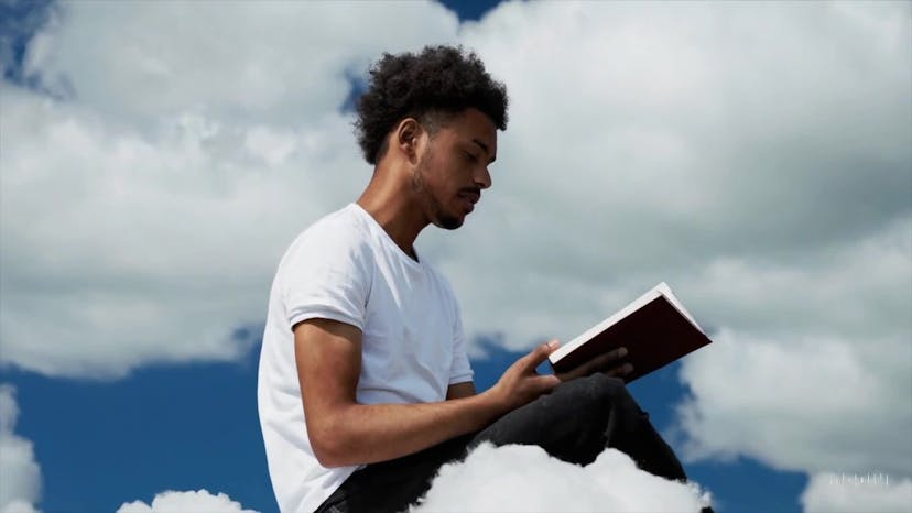 A young man at his 20s is sitting on a piece of cloud in the sky, reading a book.
